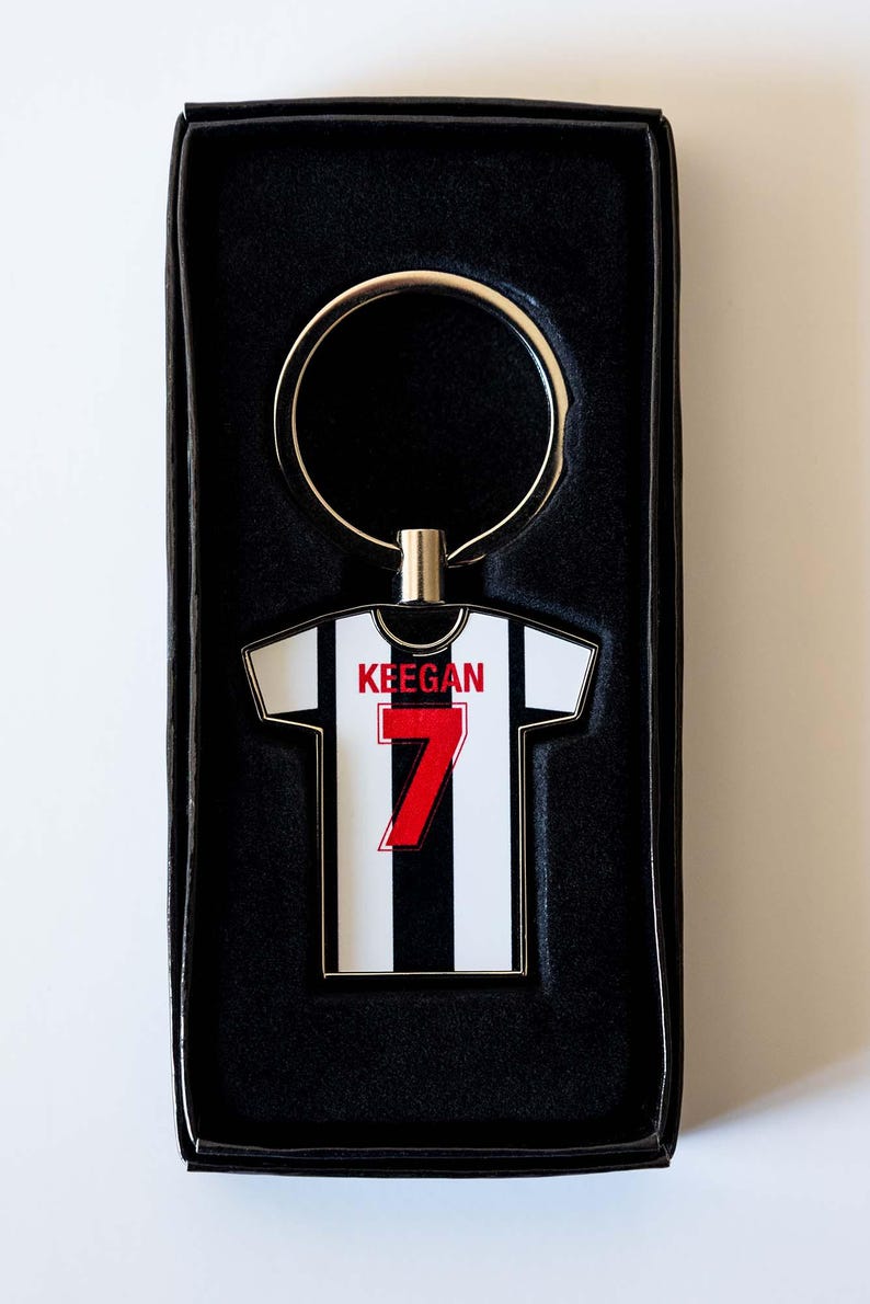 May include: A keychain in a black gift box. The keychain is shaped like a soccer jersey with black and white stripes, the number 7 in red, and the name "Keegan" above it. The keychain has a gold-colored ring.