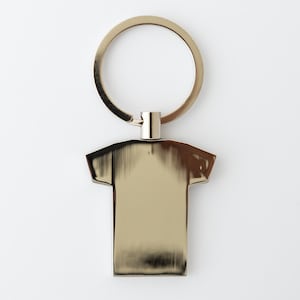 May include: A gold-colored metal keychain with a t-shirt shaped pendant. The keychain has a circular ring and a small connecting piece. The t-shirt pendant is smooth and reflective, with a brushed metal appearance.