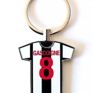 Puede incluir: Un llavero con forma de camiseta de f&uacute;tbol con el nombre "GASCOIGNE" y el n&uacute;mero "8" en rojo. La camiseta tiene un dise&ntilde;o a rayas blancas y negras, con un llavero plateado.