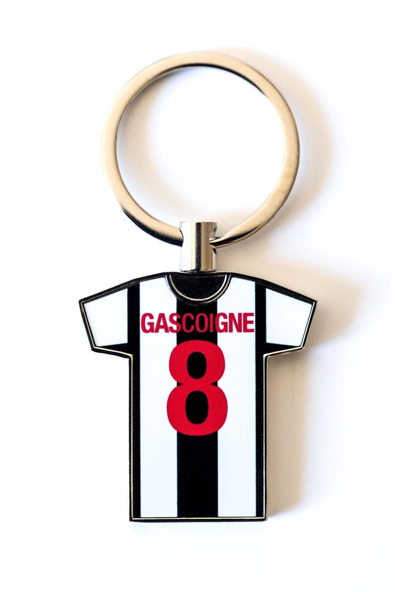Puede incluir: Un llavero con forma de camiseta de f&uacute;tbol con el nombre "GASCOIGNE" y el n&uacute;mero "8" en rojo. La camiseta tiene un dise&ntilde;o a rayas blancas y negras, con un llavero plateado.