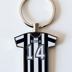 May include: A black and white striped jersey-shaped keychain with the name "GINOLA" and the number "14" printed on it. The keychain has a silver ring attached to the top.