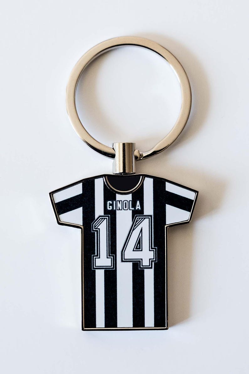 May include: A black and white striped jersey-shaped keychain with the name "GINOLA" and the number "14" printed on it. The keychain has a silver ring attached to the top.