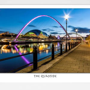 May include: A photograph of The Quayside at dusk. The image shows a bridge with a pink arch, buildings with illuminated windows, and a reflection in the water. The sky is a deep blue, and the scene is lit by streetlights. The text "THE QUAYSIDE" is at the bottom.