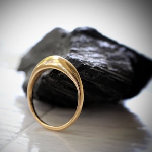 May include: A simple gold band ring with a rounded, slightly domed top. The ring is sitting on a dark grey rock.