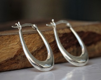 May include: A pair of polished silver hoop earrings with a smooth, elongated U-shaped design. Each earring features a hinged post closure at the top, showcasing a reflective metallic finish and a curved, tapered silhouette that narrows toward the top.