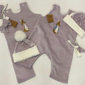 Może przedstawiać: Dwa komplety ubranek dla niemowląt z lazurowym kombinezonem z weluru z haftem Peter Rabbit, białym czapeczką z pomponem, białą opaską na głowę i lazurową opaską na głowę z kokardką.