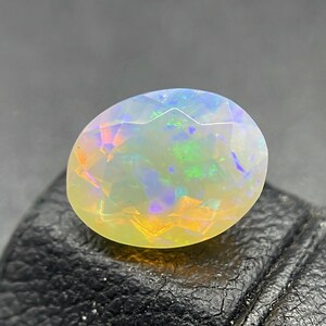 Könnte beinhalten: Ein facettierter ovaler Opal-Edelstein mit Farbspiel. Der Edelstein zeigt eine Mischung aus gelben, orangen, grünen, blauen und violetten Farbtönen. Der Stein ist vor einem dunklen Hintergrund platziert, der seine irisierenden Eigenschaften hervorhebt.