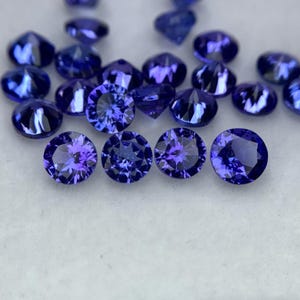 Può includere: Una collezione di pietre preziose rotonde e sfaccettate di colore viola. Le pietre preziose sono disposte in un mucchio sciolto su una superficie bianca.