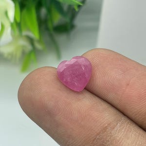 Puede incluir: Una gema facetada en forma de corazón, de un vibrante color rosa. La gema se sostiene entre los dedos, mostrando sus superficies pulidas y el juego de la luz en sus facetas. El fondo está borroso, resaltando el color y la forma de la gema.