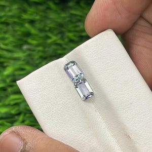 May include: Two rectangular, pale blue gemstones with a hint of purple are shown on a white surface. The gems are placed next to each other, highlighting their cut and clarity. The background is a blurred green.