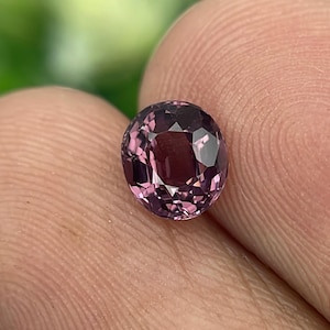 May include: A round, faceted gemstone with a deep purplish-pink hue. The gem is held between fingers, showcasing its polished surface and internal brilliance. The background is softly blurred, highlighting the gem's color and clarity.