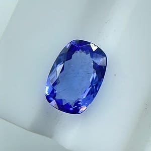 May include: A faceted, oval-shaped gemstone with a deep blue hue. The gem's surface reflects light, showcasing its multiple facets and internal structure. The stone is set against a white background, highlighting its color and clarity. This is a precious stone.