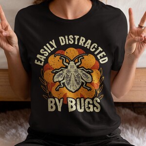 May include: Black t-shirt with the text "EASILY DISTRACTED BY BUGS" in a distressed font. The graphic features a detailed insect illustration in a circular design with orange and yellow hues. The shirt is worn by a person.