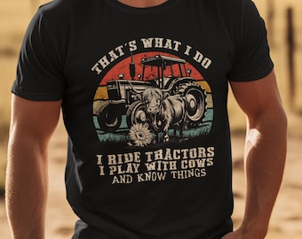 May include: A black t-shirt featuring a graphic print of a tractor and a Highland cow in front of a sunset background. The text reads: THAT'S WHAT I DO, I RIDE TRACTORS, I PLAY WITH COWS, AND KNOW THINGS. The design includes a sunflower at the bottom.
