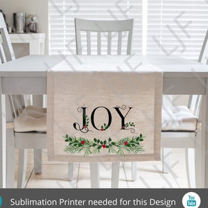 May include: A white table runner with the word "JOY" in black script with a green and red holly wreath design. The runner is on a white table with two white chairs.