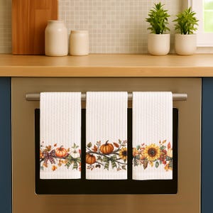 May include: Three white kitchen towels with a fall-themed design. Each towel features a decorative border with pumpkins, sunflowers, and fall leaves. The towels are hanging on a silver bar, perfect for the autumn season.