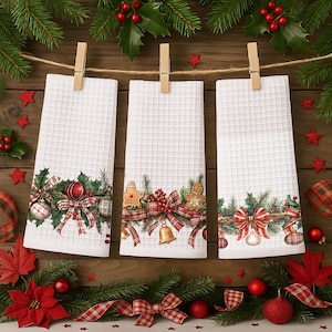 May include: Three white waffle-weave Christmas kitchen towels hanging from wooden clothespins. Each towel features a festive border with holly, ornaments, gingerbread cookies, and red plaid bows. The towels are decorated with red and green accents.