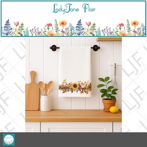 May include: White kitchen towel with a sunflower and fall leaves design. The towel hangs on a black rod above a wooden countertop. A cutting board, wooden spoons, and a small potted plant are also visible.