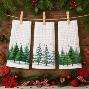 May include: Three white waffle-weave Christmas towels hanging from a string with wooden clothespins. Each towel features a winter forest scene with green and snow-covered trees. The scene is decorated with red stars, holly, and ornaments.