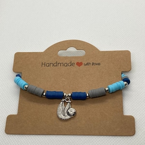May include: A handmade bracelet featuring blue, grey, and silver beads, with a silver sloth charm. The bracelet is presented on a brown card with the text "Handmade with love".