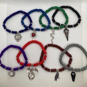 Op de afbeelding: Een verzameling kleurrijke armbanden met diverse bedels. De armbanden zijn gemaakt van platte, ronde kralen in tinten paars, rood, bruin, grijs, blauw, groen en zwart. Bedels zijn onder meer een hart met een kruis, een draak, een raaf en een kist.