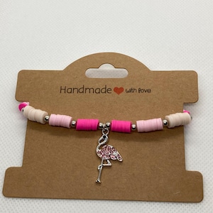 May include: A handmade bracelet with a flamingo charm. The bracelet features pink, white, and silver beads. The flamingo charm is silver with pink crystal detailing. The bracelet is displayed on a brown card with the text "Handmade with love."