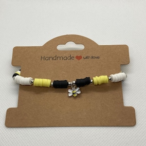 May include: Handmade bracelet featuring yellow, black, and white beads with a silver-tone bee charm. The bracelet is displayed on a brown card with the text "Handmade with love".