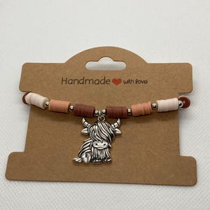 May include: A bracelet with a silver Highland cow charm, strung with brown, pink, and white beads. The bracelet is displayed on a brown card. The card has the text "Handmade with love."