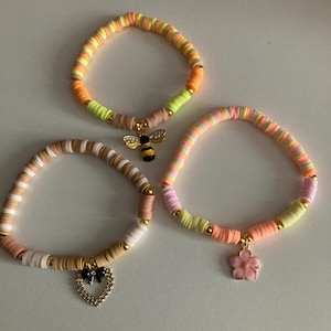 May include: Three colourful beaded bracelets. One bracelet features a gold bee charm, another a heart with a black bow, and the third a pink flower charm. The bracelets have orange, yellow, pink, and white beads.