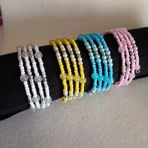 May include: Four beaded bracelets in white, yellow, turquoise, and pink. Each bracelet features multiple strands of small beads, with silver-colored accents. The bracelets are displayed on a black surface, showcasing their colors and designs.