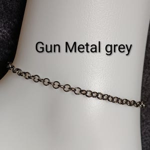 以下が含まれることがあります： ダークなガンメタルグレー仕上げの丸い連結リンクが特徴の繊細なチェーンブレスレット。チェーンの上には「Gun Metal grey」という文字が印字されており、滑らかで明るい色の湾曲した表面に対してディスプレイされています。