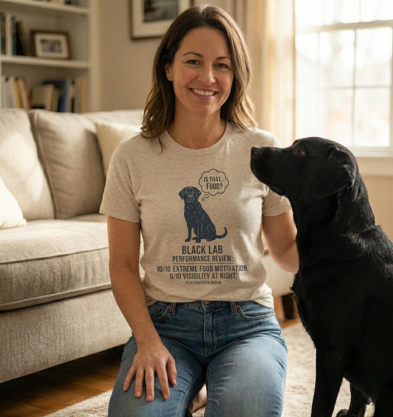 Pu&ograve; includere: T-shirt grigio chiaro con grafica di labrador nero e testo "IS THAT FOOD?" e "BLACK LAB PERFORMANCE REVIEW: 10/10 EXTREME FOOD MOTIVATION, 0/10 VISIBILITY AT NIGHT." La persona indossa jeans blu.