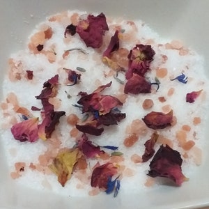 Pode incluir: Uma tigela branca cheia de sais de banho, cristais de sal rosa do Himalaia e pétalas de rosa secas. As pétalas são uma mistura de vermelho, rosa e amarelo.