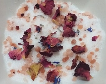 May include: A white bowl filled with bath salts, pink Himalayan salt crystals, and dried rose petals. The petals are a mix of red, pink, and yellow.