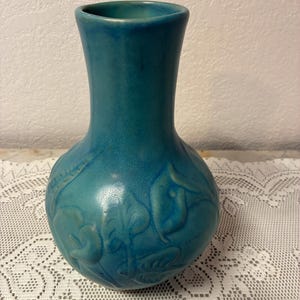 Puede incluir: Un jarrón de cerámica de color turquesa con un cuerpo bulboso y un cuello estrecho. El jarrón presenta diseños en relieve de flores y hojas alrededor del cuerpo. Se exhibe sobre un mantel de encaje blanco.