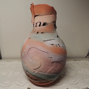 May include: A handcrafted ceramic vase with a unique, textured surface. The vase features a blend of colors, including peach, pink, and blue, with a rough, layered design. The vase is a decorative item, perfect for displaying flowers or as a standalone art piece.