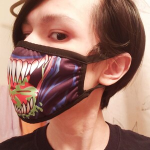 May include: A person wearing a black face mask with a colorful graphic of a monster's mouth with sharp teeth and green slime.