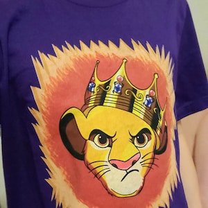 May include: Purple t-shirt with a cartoon image of Simba from The Lion King wearing a gold crown. The image is set against a background of orange flames.