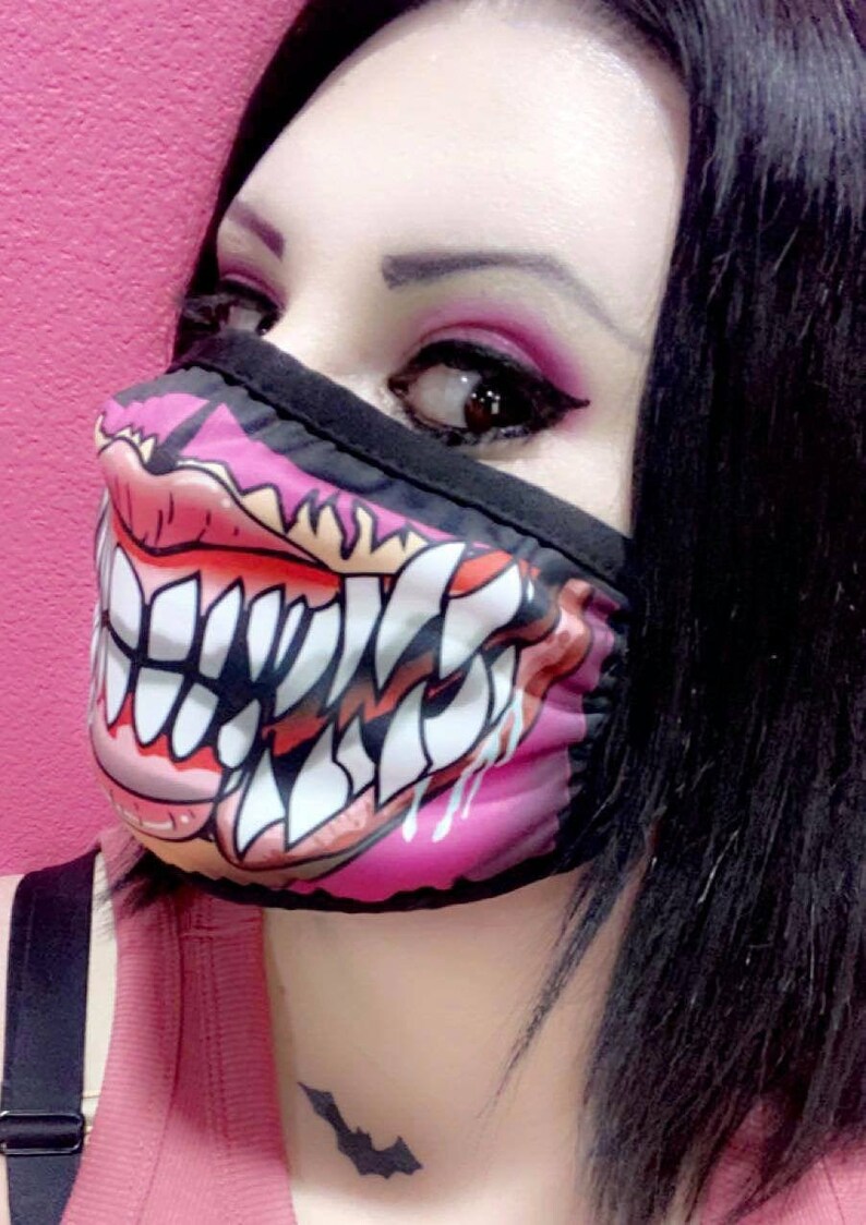 Puede incluir: Una mujer con una m&aacute;scara facial negra y rosa con un gr&aacute;fico de una boca grande y dentada. La m&aacute;scara cubre la mitad inferior de su rostro. Tiene el pelo oscuro y lleva un top rosa sin mangas. Tiene un peque&ntilde;o tatuaje de murci&eacute;lago en el cuello.