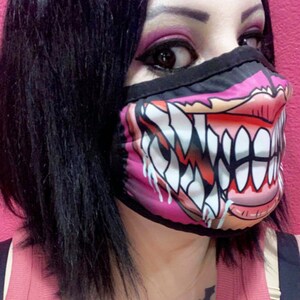 Puede incluir: Una mascarilla facial rosa y negra con un gr&aacute;fico de una boca de monstruo grande y dentada. La mascarilla tiene una banda el&aacute;stica negra para las orejas.