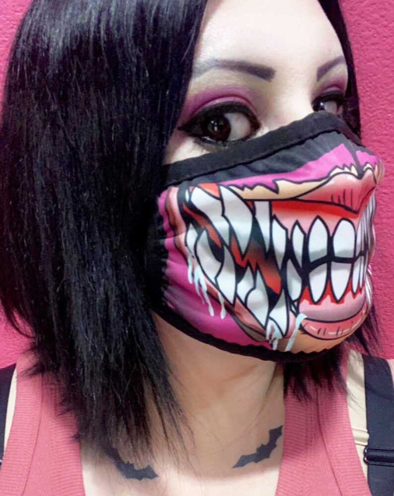 Puede incluir: Una mascarilla facial rosa y negra con un gr&aacute;fico de una boca de monstruo grande y dentada. La mascarilla tiene una banda el&aacute;stica negra para las orejas.