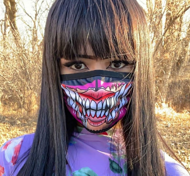 Puede incluir: Una persona lleva una m&aacute;scara facial negra con un gr&aacute;fico rosa y blanco de la boca de un monstruo con dientes afilados. La m&aacute;scara tiene un borde negro y cubre la nariz y la boca de la persona.