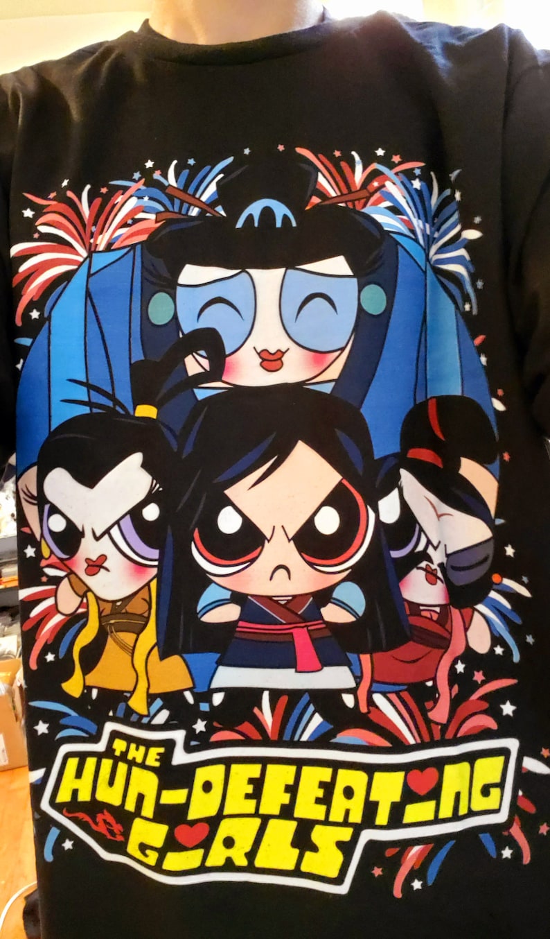 May include: Black t-shirt with a graphic of four cartoon girls in a superhero pose. The girls are wearing colorful outfits and have large eyes. The text "The Hun-Defeating Girls" is printed in yellow with a red heart. The background features red, white, and blue fireworks.