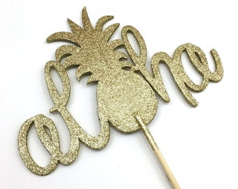 May include: Gold glitter cake topper with the word "Aloha" and a pineapple design.