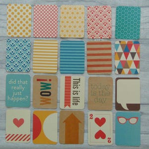 Op de afbeelding: Een verzameling kleurrijke, rechthoekige kaarten met verschillende patronen en tekst. Ontwerpen omvatten stippen, strepen, geometrische vormen en zinnen als "WOW!" en "This is life." De kaarten zijn gerangschikt op een licht houtnerfoppervlak.