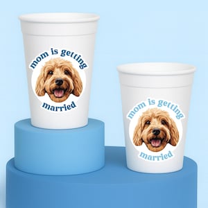 May include: Two white plastic cups featuring a circular graphic of a golden-brown dog face with the blue text "mom is getting married" arched around the image. The cups are smooth, tapered, and have a simple rim at the top edge of each vessel.