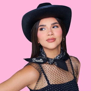 May include: A woman wearing a black cowboy hat with a black bandana around her neck. The bandana has the word "Brielle" embroidered on it. She is wearing a black mesh top with a black and white pattern.