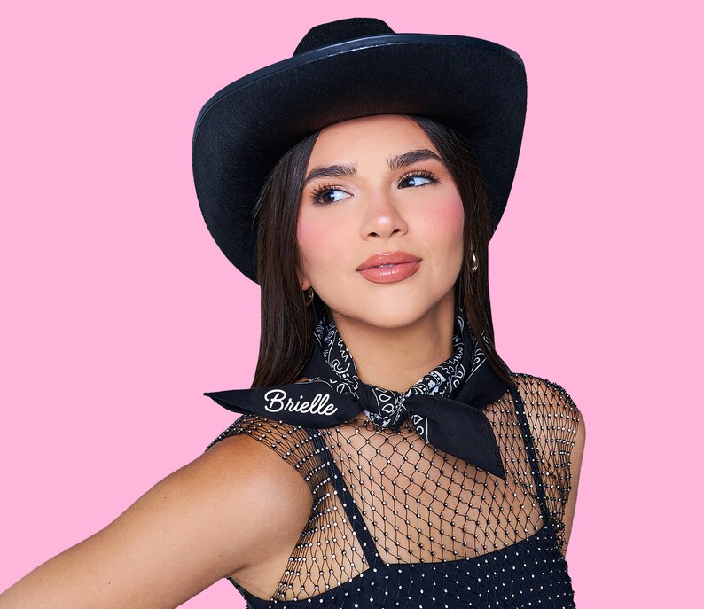 May include: A woman wearing a black cowboy hat with a black bandana around her neck. The bandana has the word "Brielle" embroidered on it. She is wearing a black mesh top with a black and white pattern.