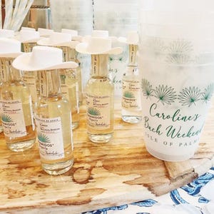 May include: A collection of small tequila bottles with white cowboy hat toppers, arranged on a wooden surface. A large plastic cup with text that reads "Caroline's Bach Weekend Isle of Palm" is also visible.