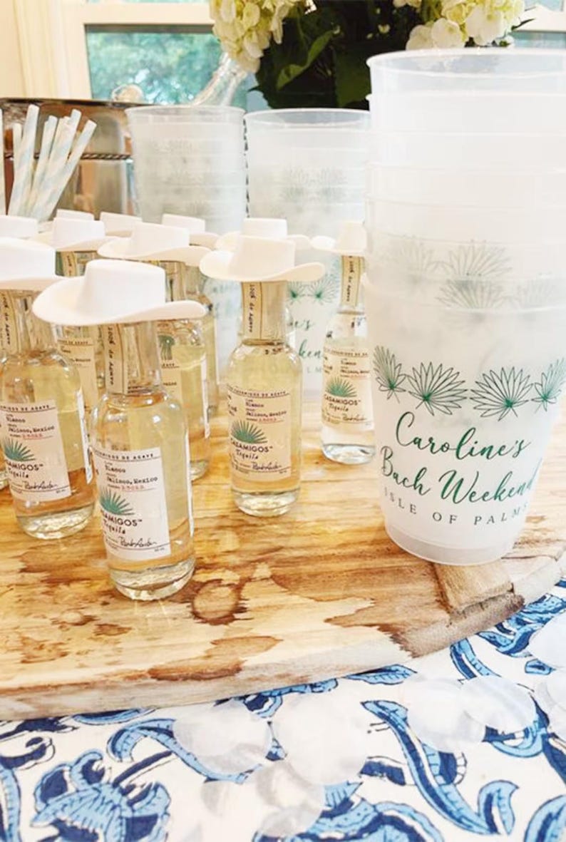May include: A collection of small tequila bottles with white cowboy hat toppers, arranged on a wooden surface. A large plastic cup with text that reads "Caroline's Bach Weekend Isle of Palm" is also visible.